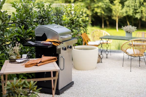 Gasgrill auf einer Kiesterrasse im Garten, dahinter Gartenmöbel