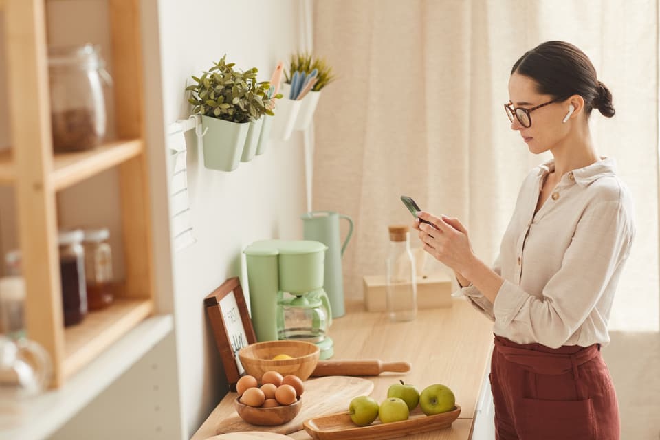 Frau in einer hellen Küche schaut auf ihr Smartphone, neben ihr eine salbeigrüne Filterkaffeemaschine und eine Kanne